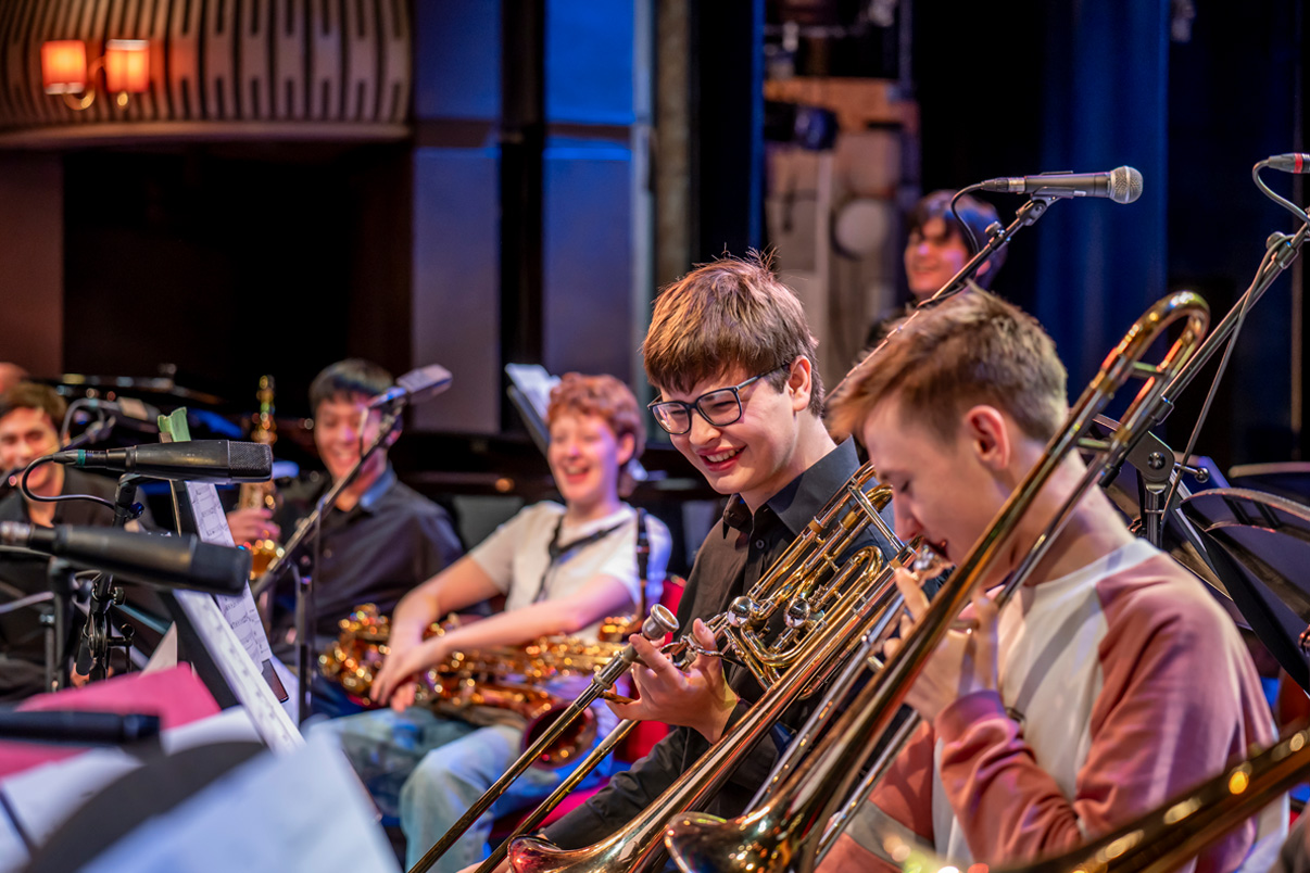Students sitting in a rehearsal, holding their trombones, smiling and chatting together.
